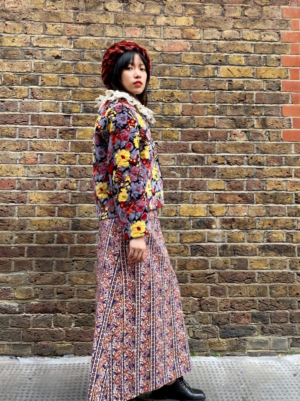 Model wearing late 1960s paisley maxi dress with square neckline and flared skirt in burnt orange and navy with a floral jacket