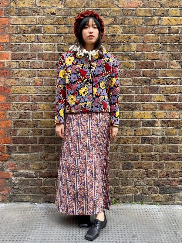 Model wearing late 1960s paisley maxi dress with square neckline and flared skirt in burnt orange and navy with a floral jacket